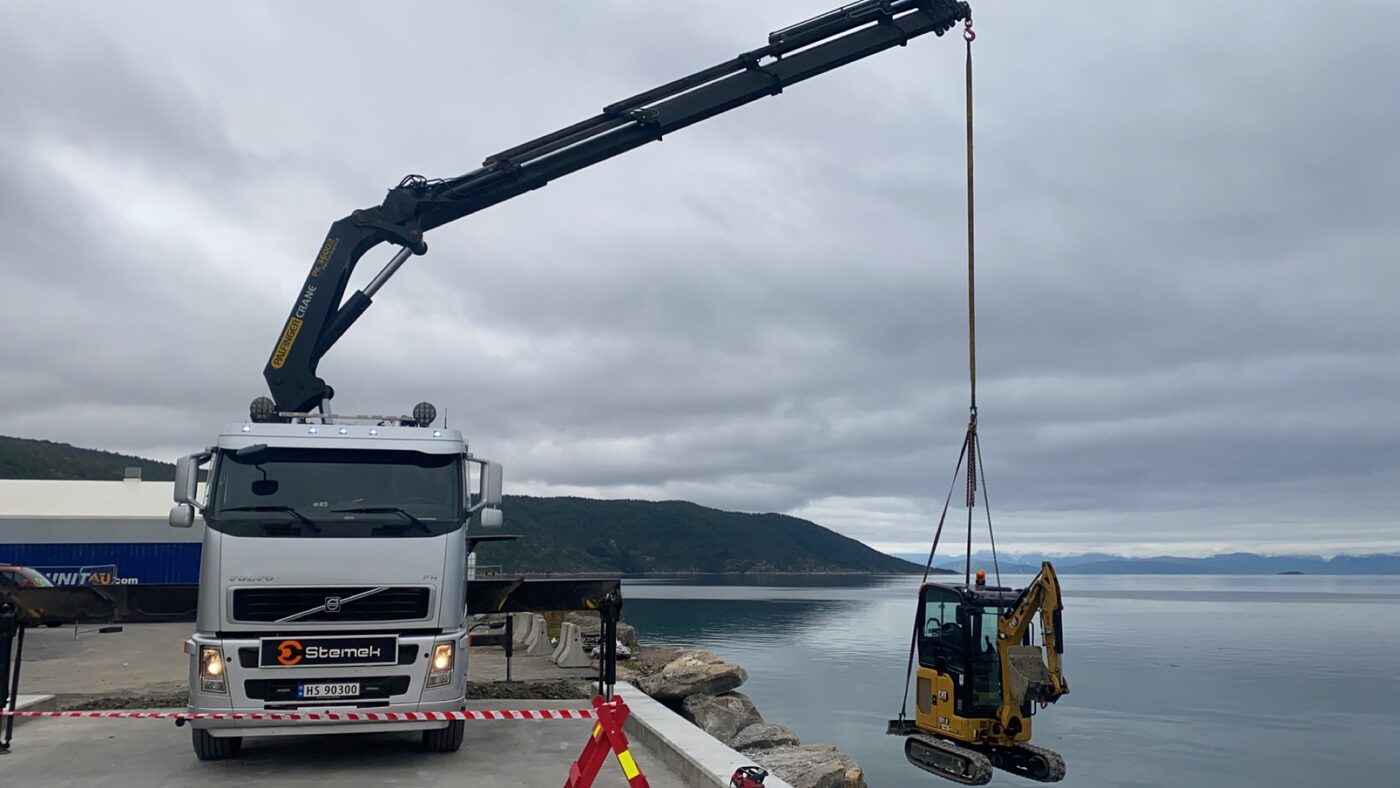 Stemek-kranbil løfter gravemaskin med fjord i bakgrunnen